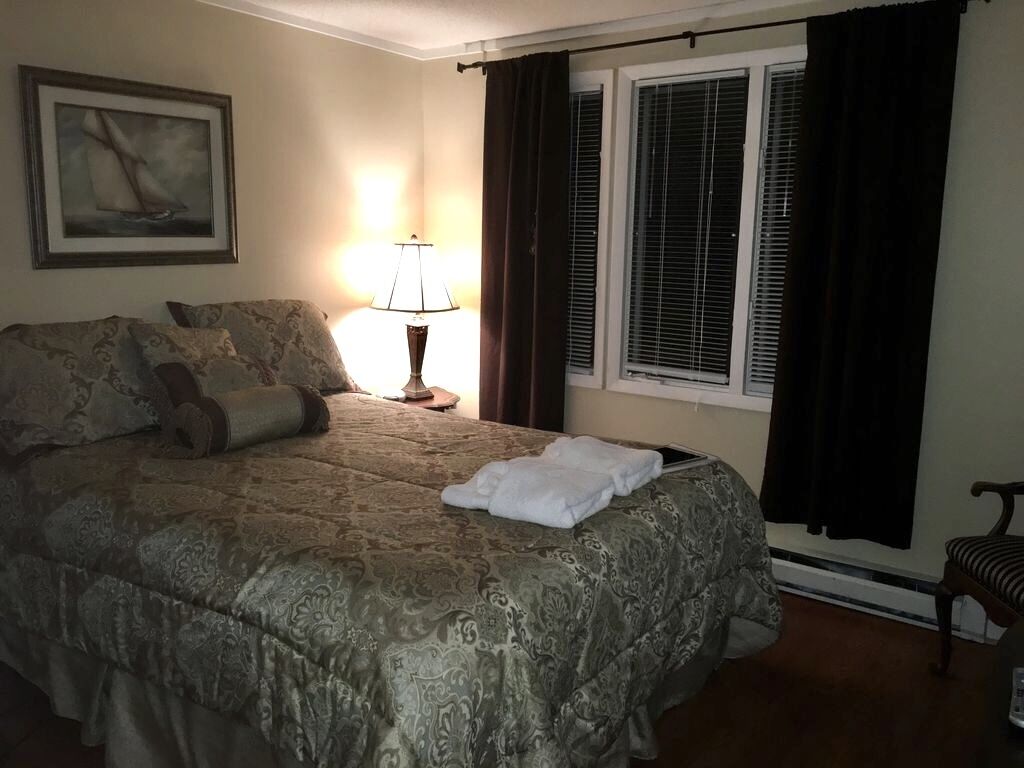 A neatly made bed with a patterned bedspread, a bedside lamp, a framed picture on the wall, and a window with blinds.