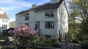 Une maison à deux étages avec un porche à l’avant, un petit jardin avec des fleurs roses et une clôture en bois.