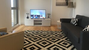 Ein modernes Wohnzimmer mit einem Flachbildfernseher auf einem weißen Schrank, einem dunklen Sofa, einem gemusterten Teppich und einem kleinen Beistelltisch mit zwei Blumentöpfen.