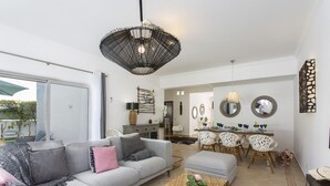 Een moderne woonkamer met een grijze bank, een geweven salontafel, en een eetgedeelte met witte stoelen en een tafel.
