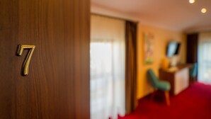 Quarto de hotel com porta de madeira marcada com o número 7, tapete vermelho, escrivaninha com um computador e uma televisão na parede.