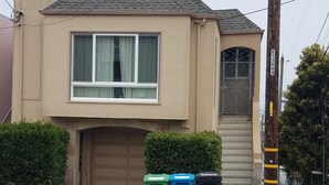 A two-story house with a beige facade, a garage door, and a small front yard with three trash bins. - Beautiful House with Panoramic SF Bay Views (San Francisco)