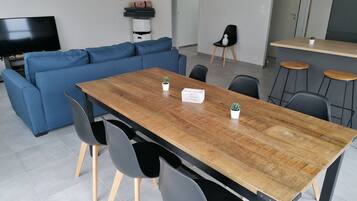 Un salon moderne avec un canapé bleu, une table à manger en bois et des chaises noires. On aperçoit une petite plante en pot sur la table et un téléviseur au fond de la pièce.