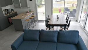A modern kitchen with a wooden countertop and bar stools, a dining area with a wooden table and chairs, and a blue sofa in the foreground. - New house 350m from the beach in Cléder. Rated 3 stars. (Cléder)