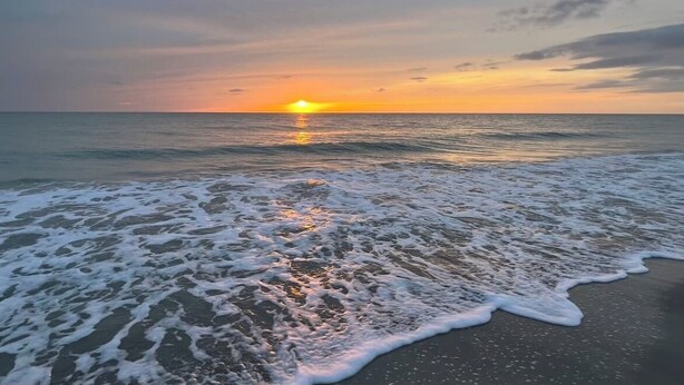 Een strandscène met golven die de kust naderen en een zonsondergang op de achtergrond.
