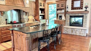 A kitchen with a large island, wooden cabinets, a stone fireplace, and a view of trees through a window.