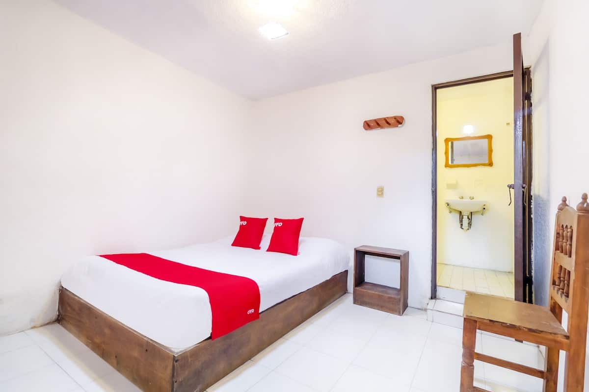 A bedroom with a wooden bed, a small bedside table, a chair, a door leading to a bathroom, and a white wall with a light fixture.