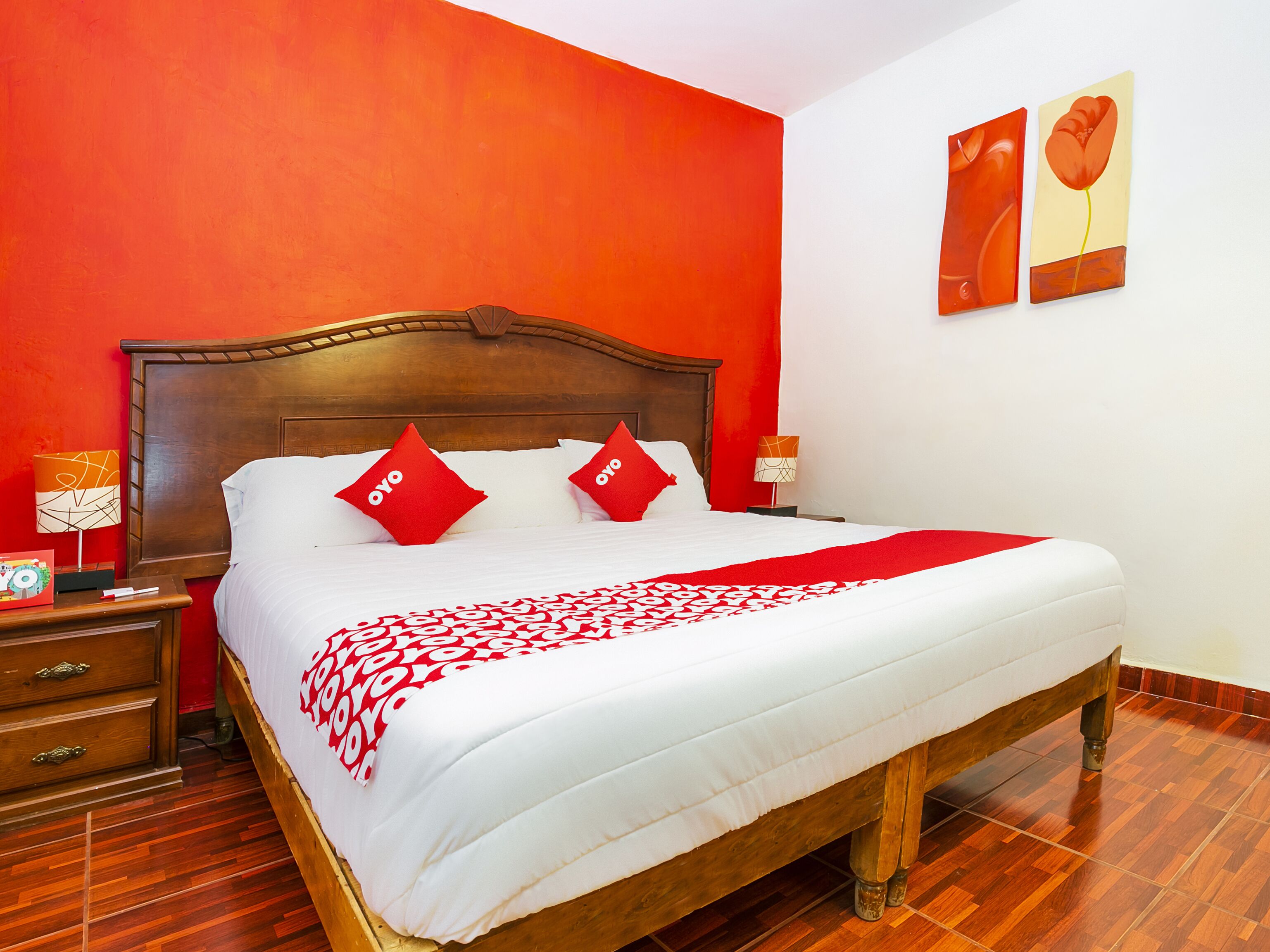 A hotel room with a wooden bed, red pillows, a white bedspread, and a red and white patterned throw.