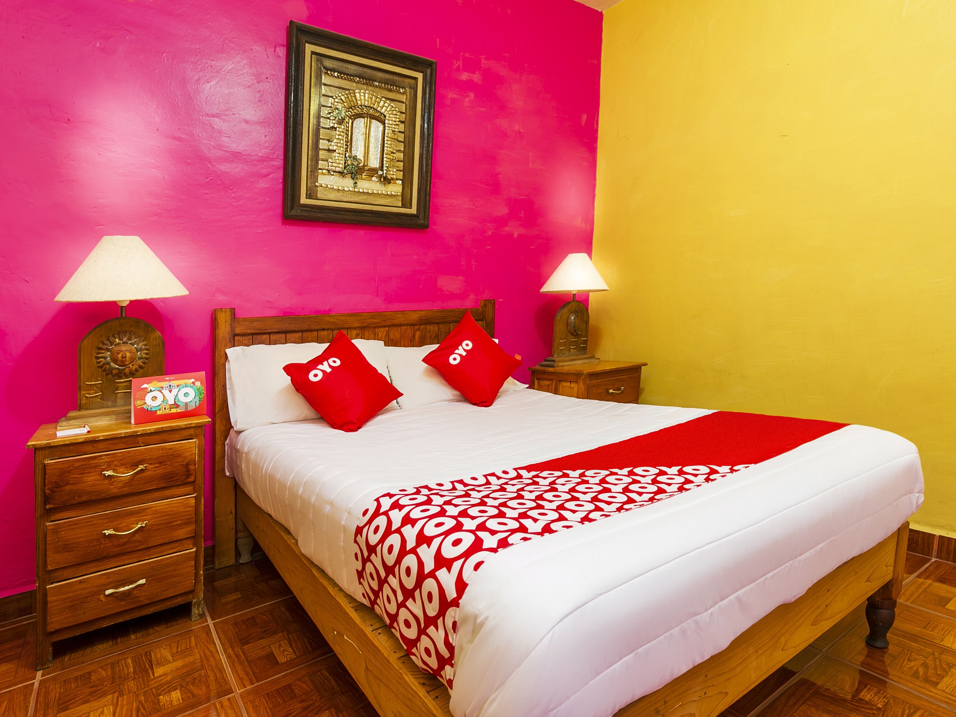 A bed with a red and white patterned bedspread, two red pillows, wooden nightstands with lamps, a framed picture on the wall, and a wooden headboard.