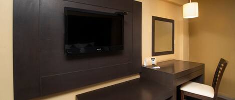 Quarto moderno com televisão de tela plana fixada em parede escura, uma mesa pequena com uma cadeira e uma luminária pendente.