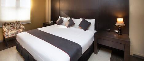 Quarto de hotel com uma cama grande, uma mesa de cabeceira, uma luminária, uma cadeira e uma janela com persianas.