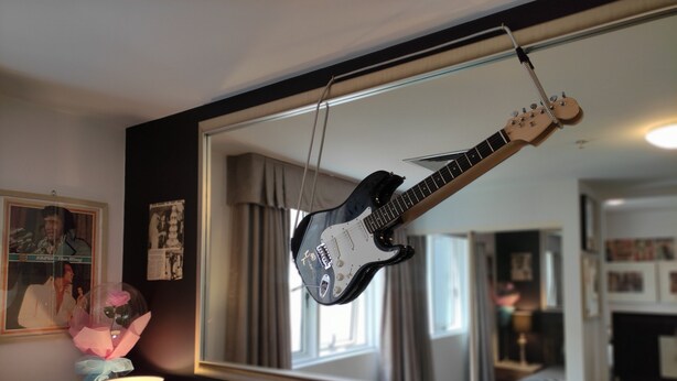 Une guitare accrochée au mur, une lampe et un miroir qui reflète une pièce.