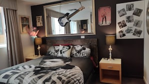 Une chambre à coucher avec un lit agrémenté d’une literie aux motifs d’Elvis Presley, une guitare accrochée au-dessus du lit et un miroir qui reflète la pièce.