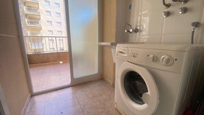 Ein Waschraum mit Waschmaschine, Warmwasserbereiter und Blick auf ein Gebäude durch eine Glastür.