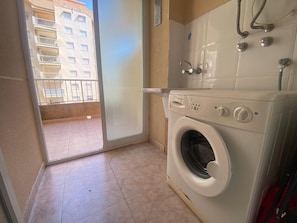 Une buanderie équipée d’un lave-linge, d’un chauffe-eau et offrant une vue sur un immeuble à travers une porte vitrée.