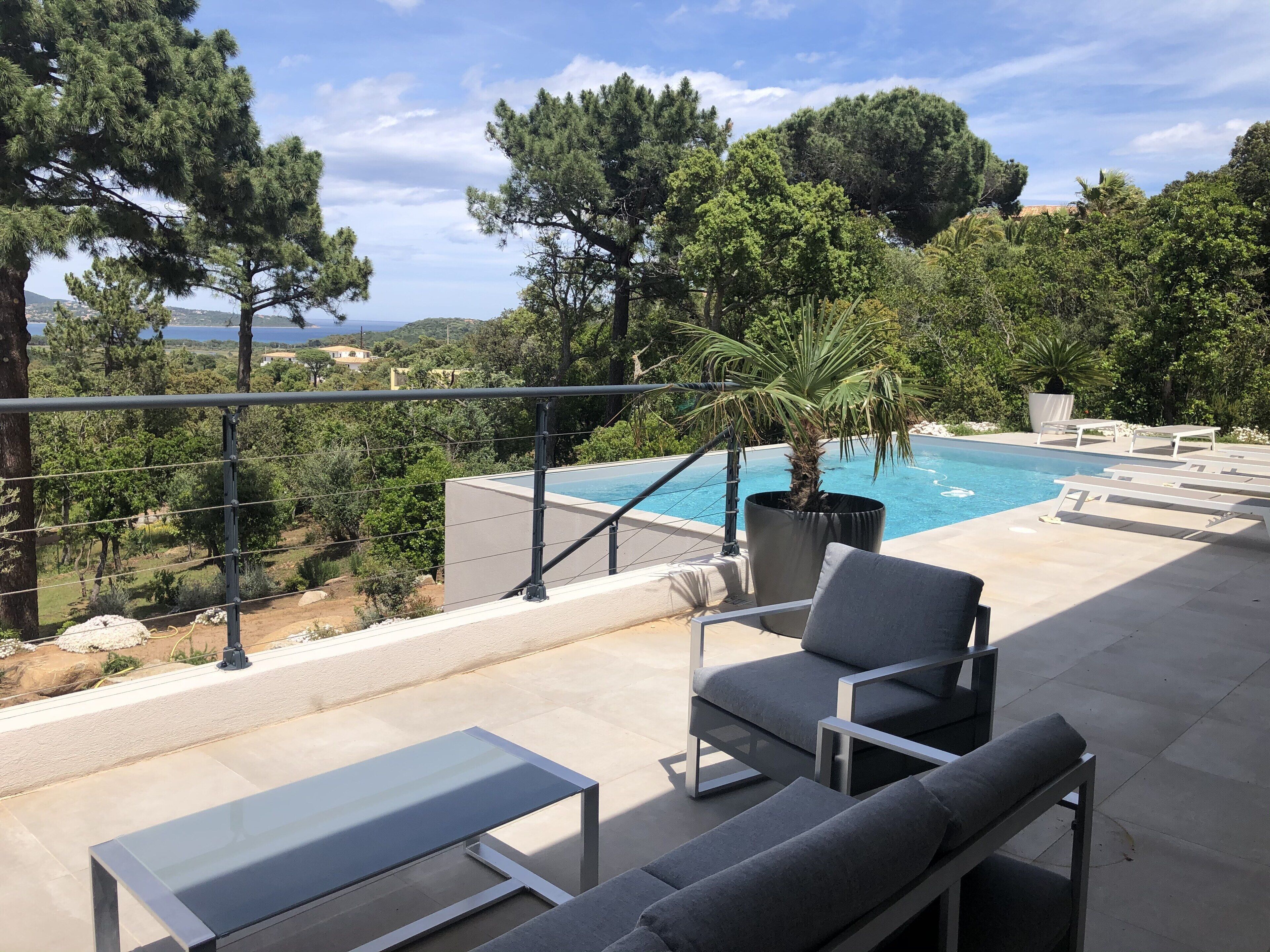 A poolside area with lounge chairs, a glass table, and a view of a pool surrounded by trees.