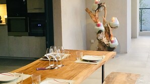 Een moderne eetruimte met een lange houten tafel gedekt voor een maaltijd, een decoratieve tak met kaarsen en een minimalistische keuken op de achtergrond.
