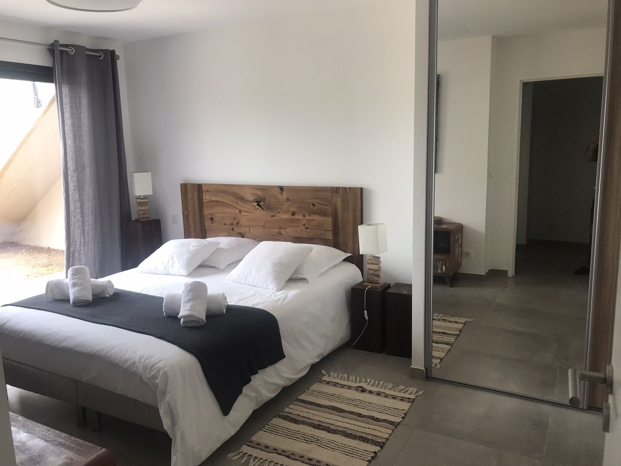 A modern bedroom with a wooden headboard, white bedding, a bedside lamp, and a view of a balcony.