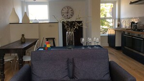 Ein modernes Wohnzimmer mit einer Couch, einem Esstisch mit bunten Bauklötzen und einer Vase mit Zweigen. Es gibt eine Küchenzeile mit Herd, Spüle und Fenster.