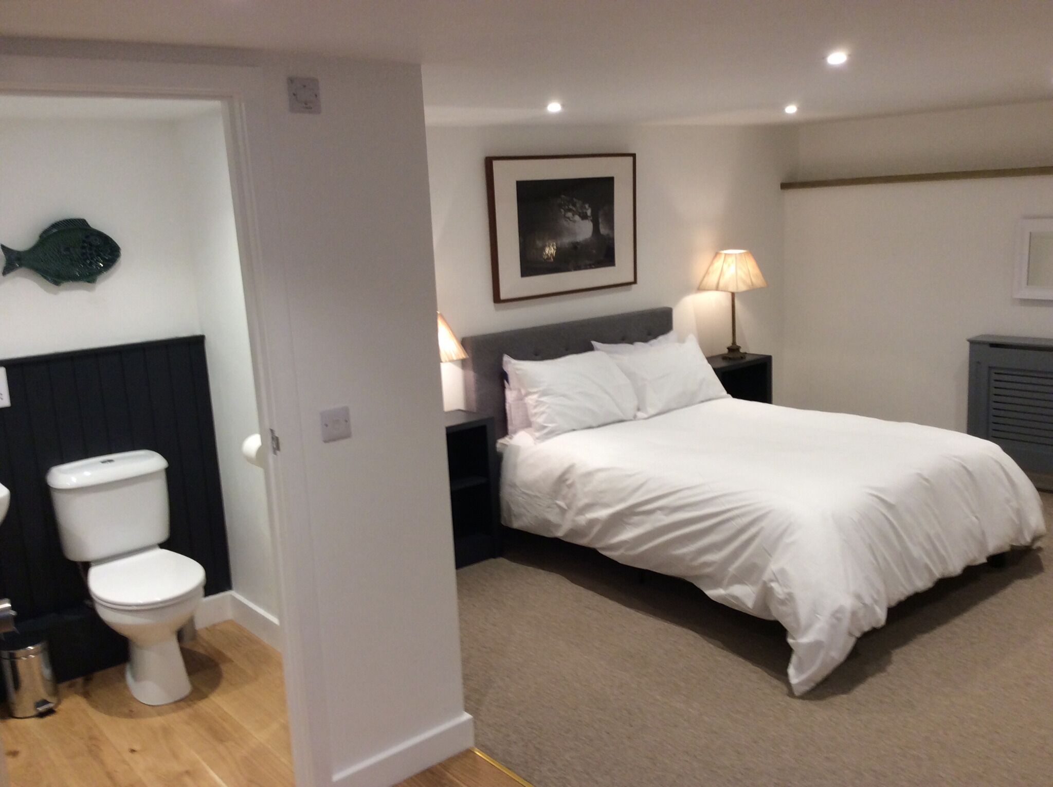A bedroom with a bed, a bedside table with a lamp, a framed picture on the wall, and a toilet in the adjacent bathroom.