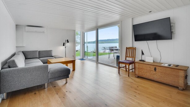 Un soggiorno moderno con un divano, un tavolo in legno, una TV a parete e vista su un lago attraverso ampie vetrate.