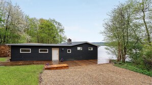 Una moderna casa nera con un patio in legno, un vialetto sterrato e vista su un lago circondato da alberi.