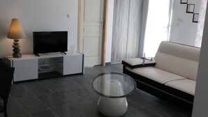 Un salon moderne avec un téléviseur à écran plat posé sur un meuble blanc, une table basse en verre, un canapé beige et une lampe.