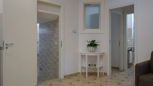 En gang med flisegulv, en hvid dør, en lille hvid hylde med en potteplante, og et rammet spejl på væggen.