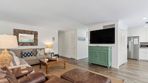 A living room with a sofa, coffee table, and ottoman, a kitchen with a TV, and a dining area with a table and chairs. - East Beach Cottage with Private Pool | Walk to Beach | Sleeps 9 (Saint Simons Island)