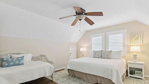 A bedroom with two beds, a ceiling fan, a window with blinds, a nightstand with a lamp, and a wall-mounted picture. - East Beach Cottage with Private Pool | Walk to Beach | Sleeps 9 (Saint Simons Island)