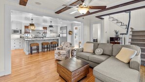 A spacious living area with a wooden coffee table, a gray sofa, and a patterned armchair. A kitchen with white cabinets and a central island is visible in the background. - Historic home in seaside village of Swansboro w/ private beach parking  (Swansboro)