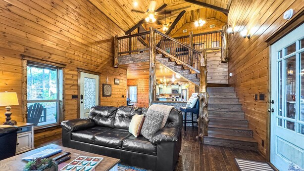 A cozy log cabin interior with a leather sofa, a dining area, and a staircase leading to an upper level. - The Good Stuff - Zipline, Gameroom, Hot Tub, & Firepit. 8 Beds / Sleeps 12 (Broken Bow)