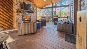 A covered outdoor area with a stone fireplace, wicker furniture, and a television mounted on the wall. - The Good Stuff - Zipline, Gameroom, Hot Tub, & Firepit. 8 Beds / Sleeps 12 (Broken Bow)