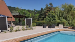 Une piscine avec une terrasse en bois, des chaises longues et un espace salon couvert offrant une vue sur une colline boisée.