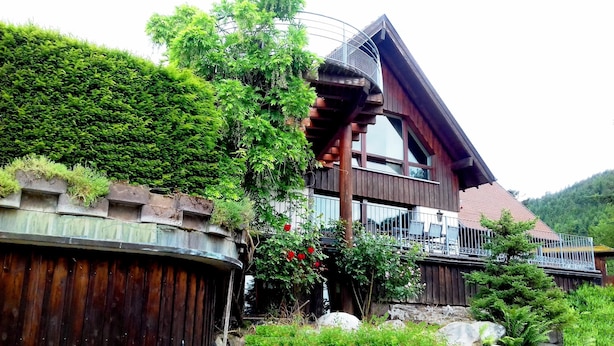 Un chalet en bois doté d’un balcon, entouré d’une végétation luxuriante et d’un mur en pierre.