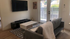 Een woonkamer met een grijze bank, een witte salontafel, een flatscreen televisie aan de muur en een schuifdeur naar buiten.