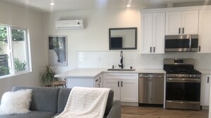 Een moderne keuken met witte kasten, een roestvrijstalen fornuis en een magnetron. In de woonkamer staat een grijze bank met een witte plaid.