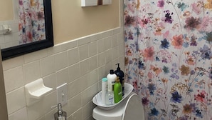 Un baño con un lavamanos blanco, un inodoro, una cortina de ducha con estampado floral, un espejo con un cuadro enmarcado y artículos de aseo en el lavamanos.