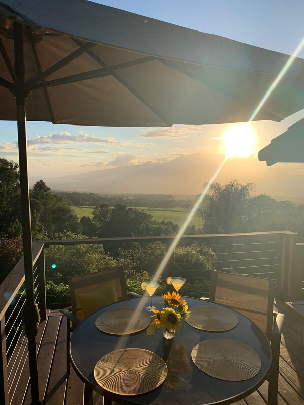 A table with four woven placemats, a vase with sunflowers, and two martini glasses with lemon slices on a balcony overlooking a scenic landscape during sunset.
