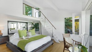 A modern bedroom with a large bed, a wooden floor, a staircase, a dining area with a table and chairs, and a view of greenery outside. - Rustic yet modern Upcountry retreat with breathtaking views! (Makawao)