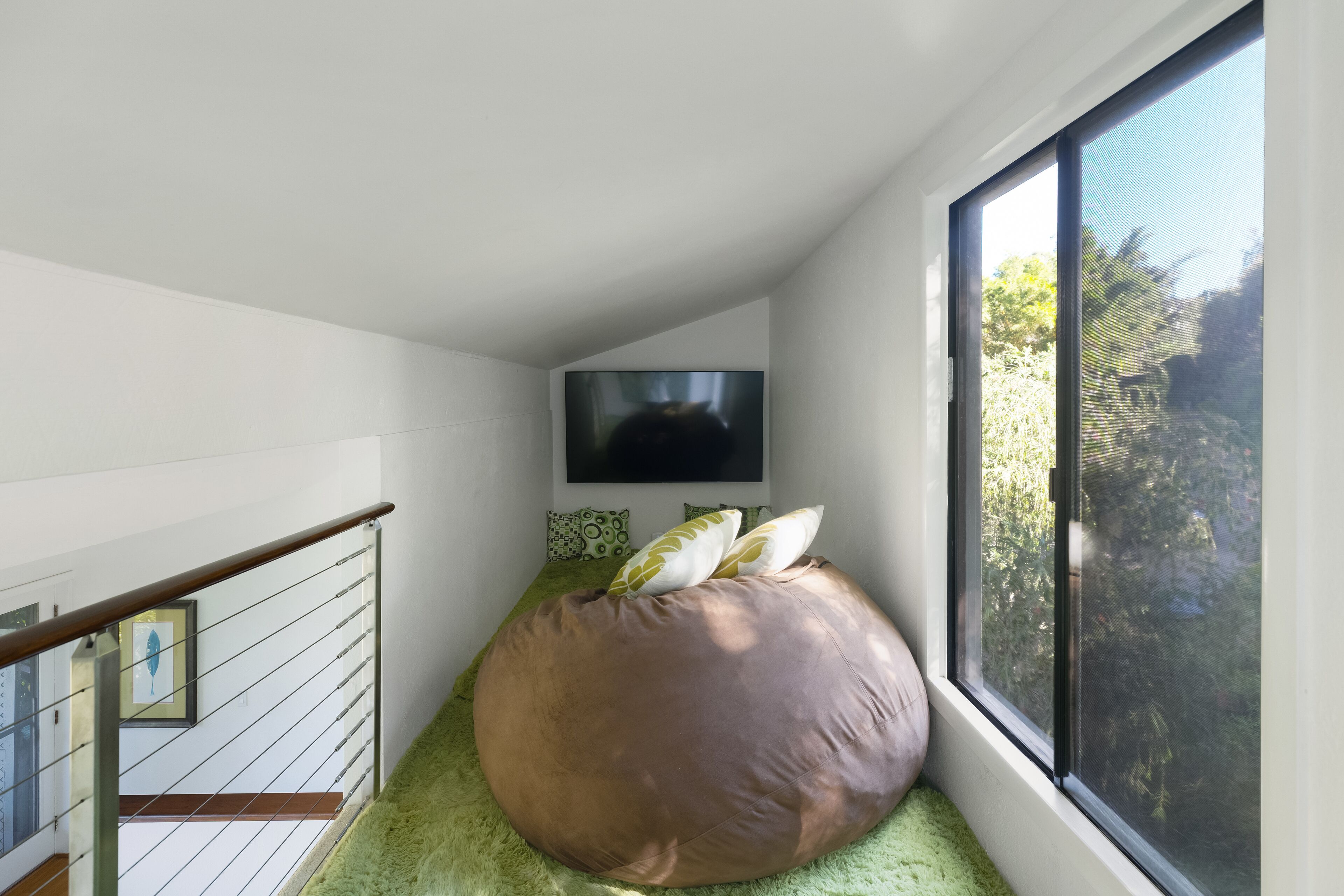 Una habitación moderna con un sillón de peluche grande, un televisor de pantalla plana montado en la pared y un ventanal con vistas a la vegetación.