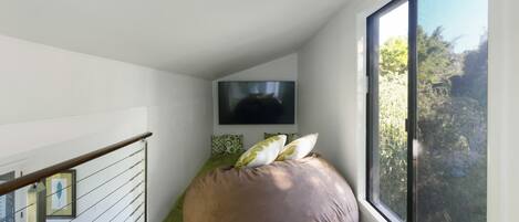 Una habitación moderna con un sillón de peluche grande, un televisor de pantalla plana montado en la pared y un ventanal con vistas a la vegetación.