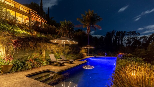 A swimming pool with a night-time view, surrounded by lush vegetation and palm trees. - Rustic yet modern Upcountry retreat with breathtaking views! (Makawao)
