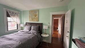 A bedroom with a bed, bedside lamps, a window with curtains, a painting on the wall, and a hallway leading to another room. - The Pistachio Room at TPH - Boston (Boston)
