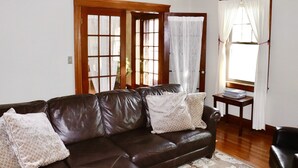 A living room with a brown leather sofa, two patterned throw pillows, a small wooden side table with a stack of books, and a window with white curtains. - The Sunset Room at TPH - Boston (Boston)