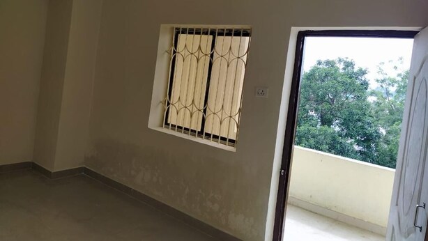 A room with a window featuring a metal grille, a door leading outside, and a balcony with a view of trees.