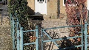 A two-story stone house with a white door and windows, a satellite dish, and a small garden with a metal gate.