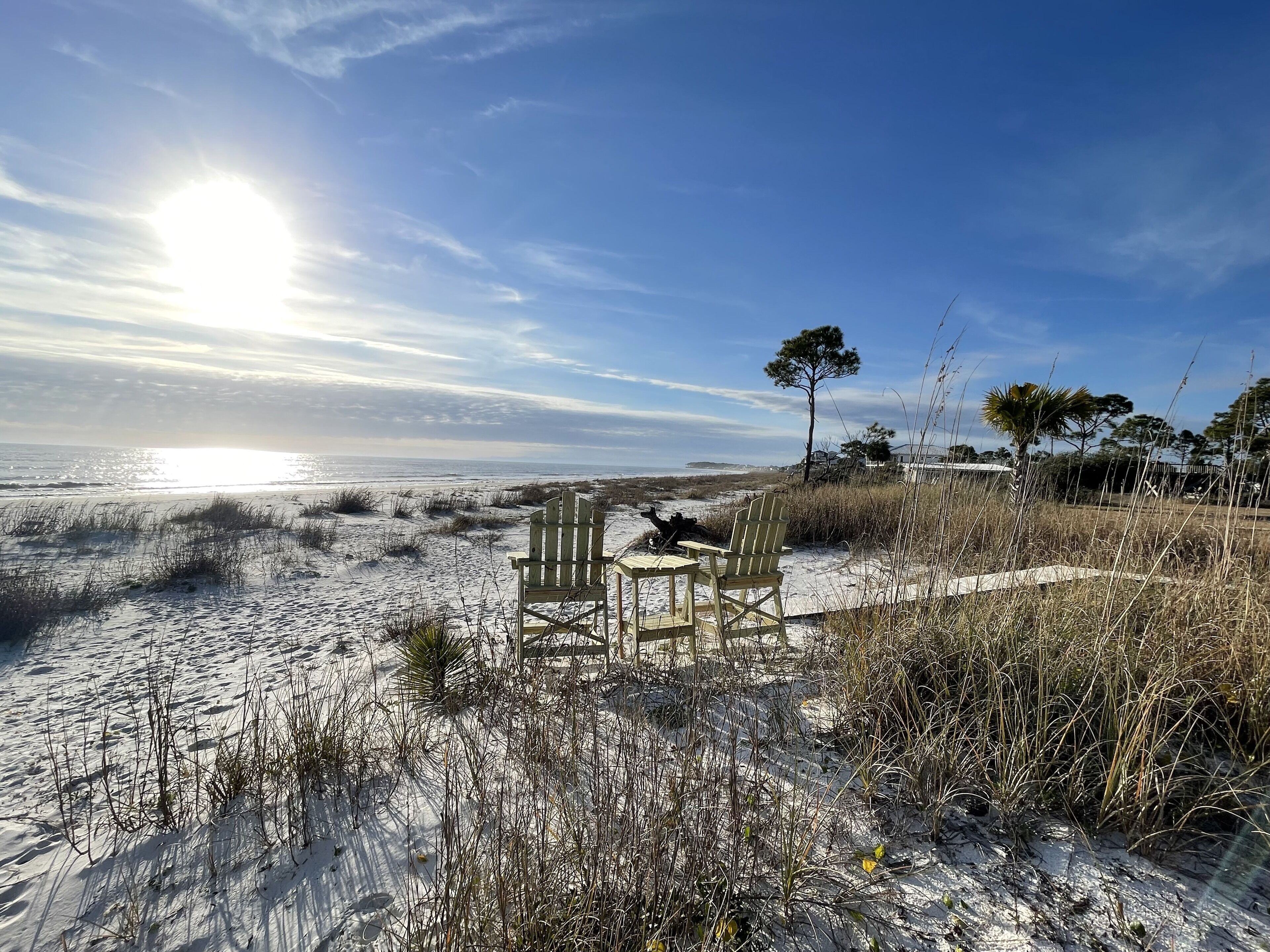 A beach scene with two wooden chairs and a table, surrounded by sand and sparse vegetation.