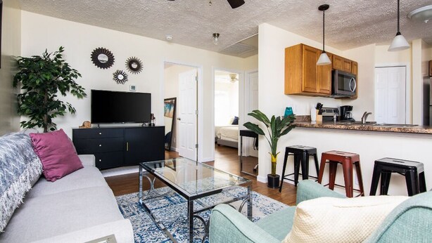 Una sala de estar moderna con un sofá, una mesa de centro y un televisor. Cuenta con una zona de cocina con un microondas y taburetes.