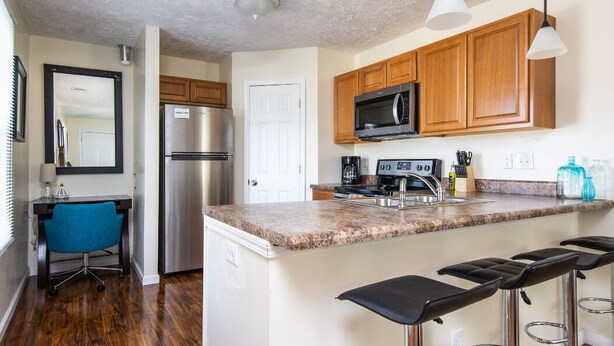 A modern kitchen with wooden cabinets, a stainless steel refrigerator, a microwave, and a granite countertop. There are three black bar stools and a blue office chair. - Sanitized Spacious East Nash Apt!  Walk to Top Restaurants! (Nashville)